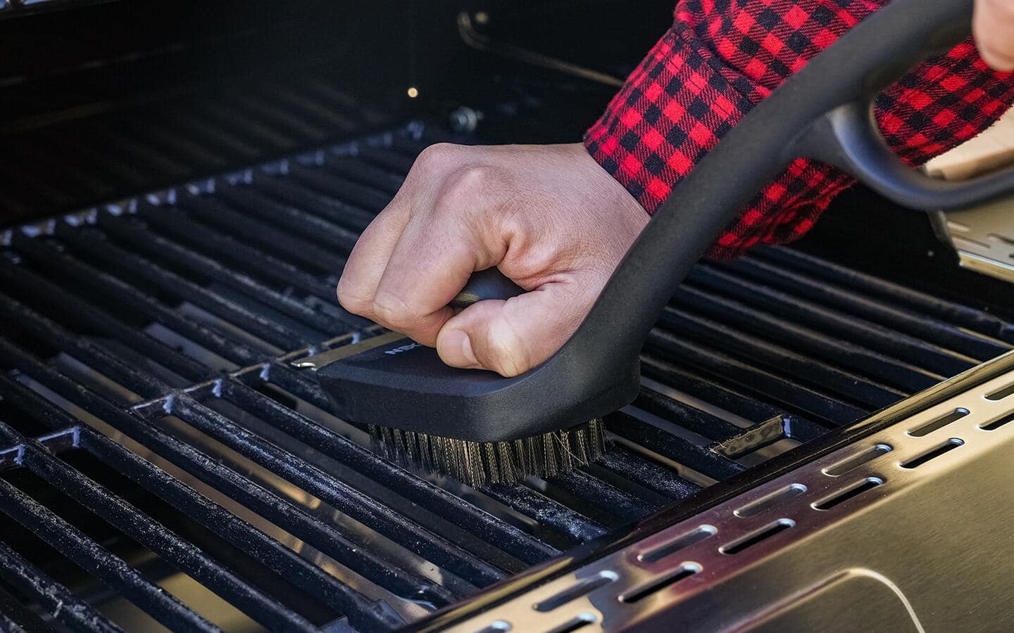 Someone in a red-checkered shirt cleaning a smoker's grill grates with a brush.