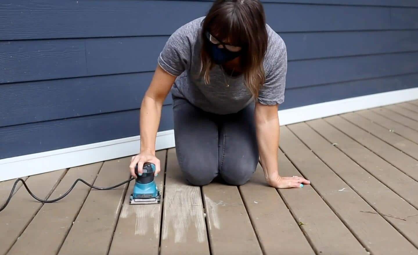 A woman uses a corded quarter-sheet sander to sand the surface of a deck during a refinishing project.