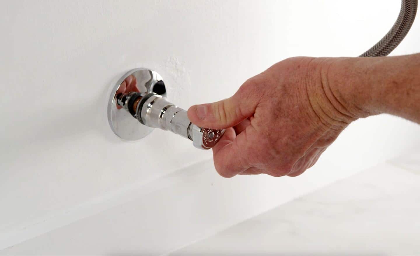 A person's left hand turning off the water supply valve to a toilet so it can be repaired.
