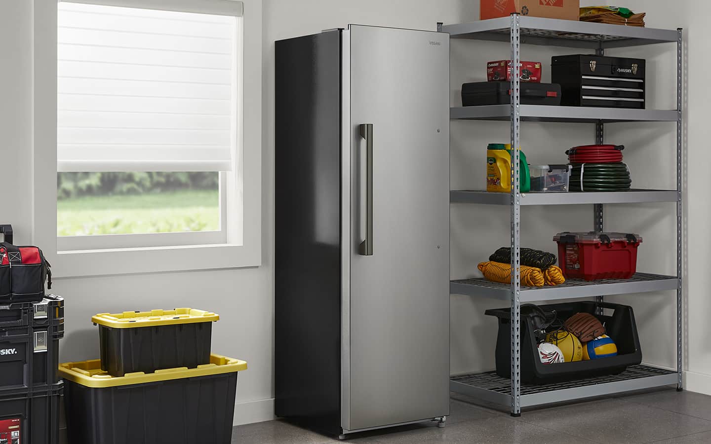 A stainless steel freezer stands in a garage next to plastic storage boxes and a black-and-yellow shelving unit.