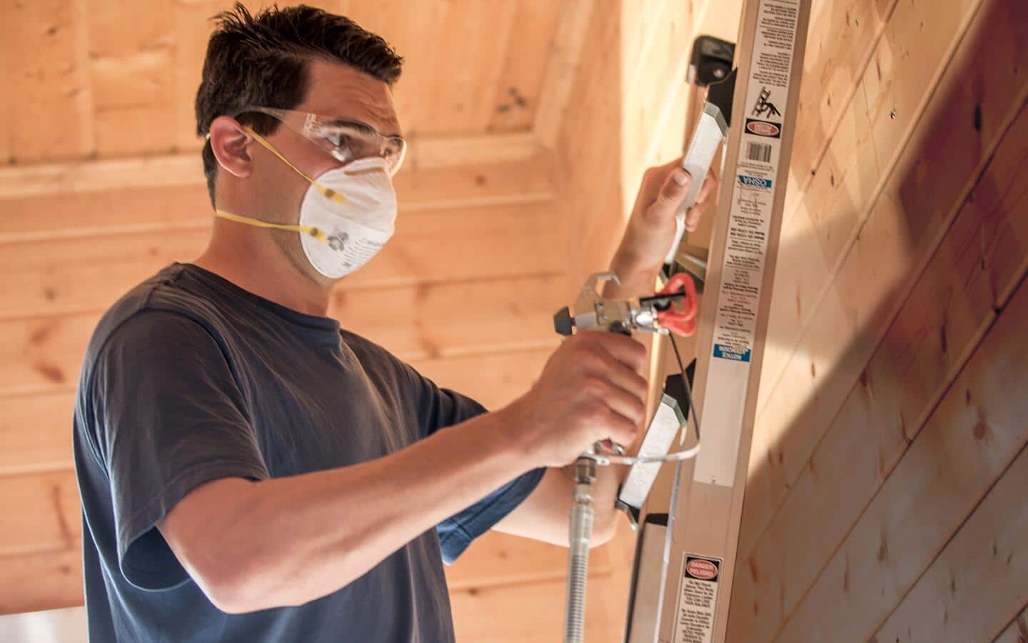 A person on a ladder in a safety mask and protective eyewear uses a paint sprayer on a wall.