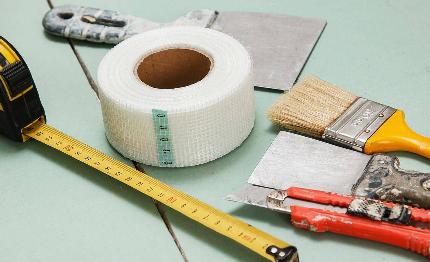 Fiberglass mesh tape, utility knives and a tape measure placed on a sheet of drywall.