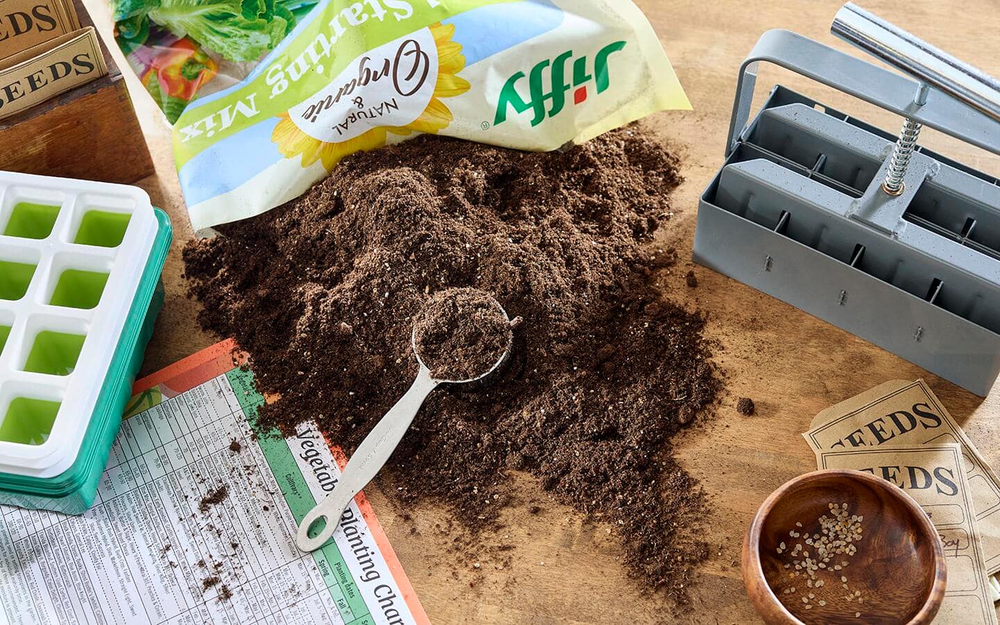 A scoop in an open bag of seed starting mix on a table with a soil block maker, a seed starting calendar and seed starting supplies