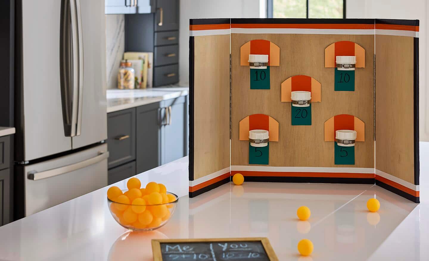 A basketball pong game sitting on a table next to a bowl of table tennis balls. A basketball pong game sitting on a table next to a bowl of table tennis balls.