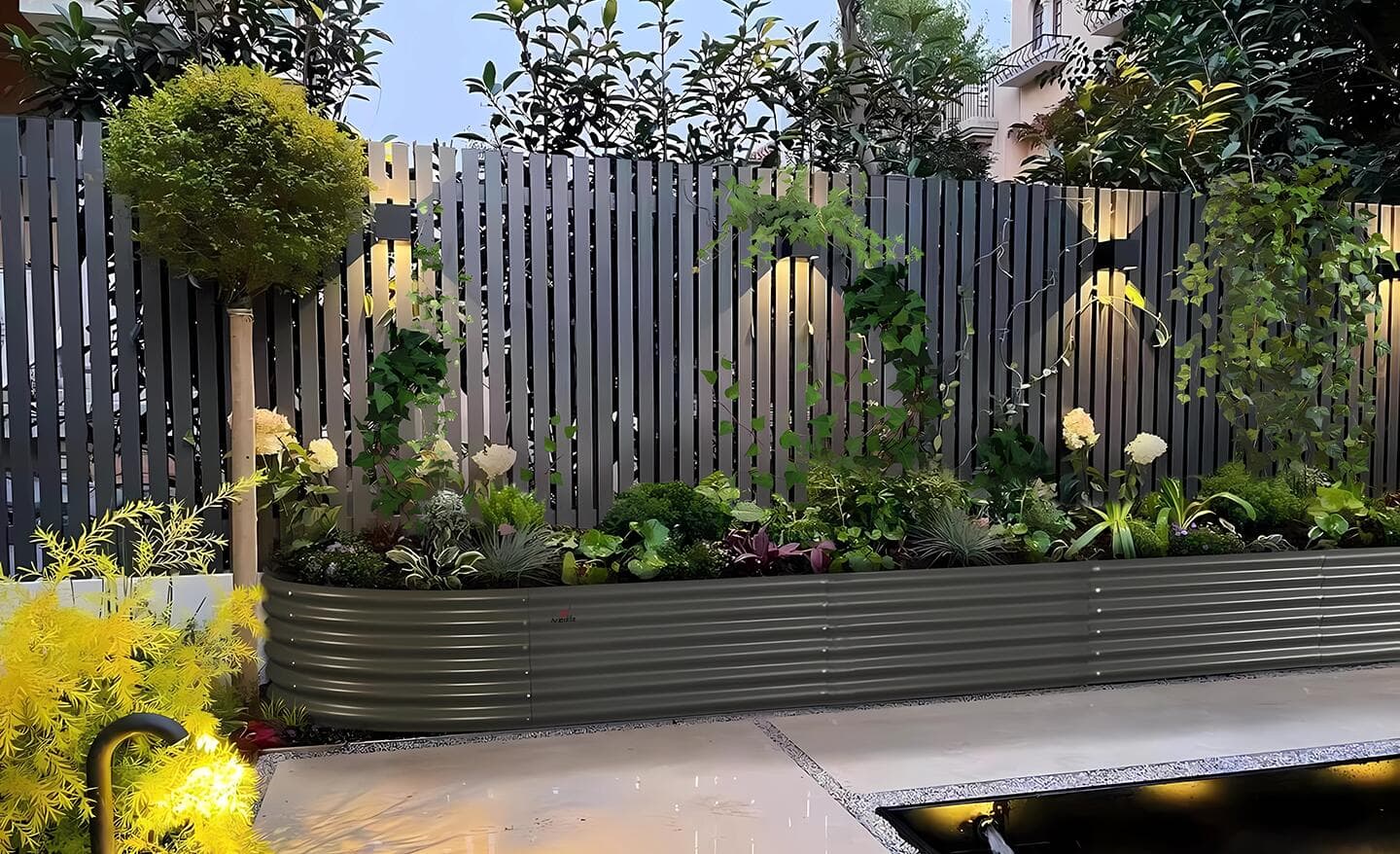 Metal raised garden bed full of greenery lined up against a fence. 