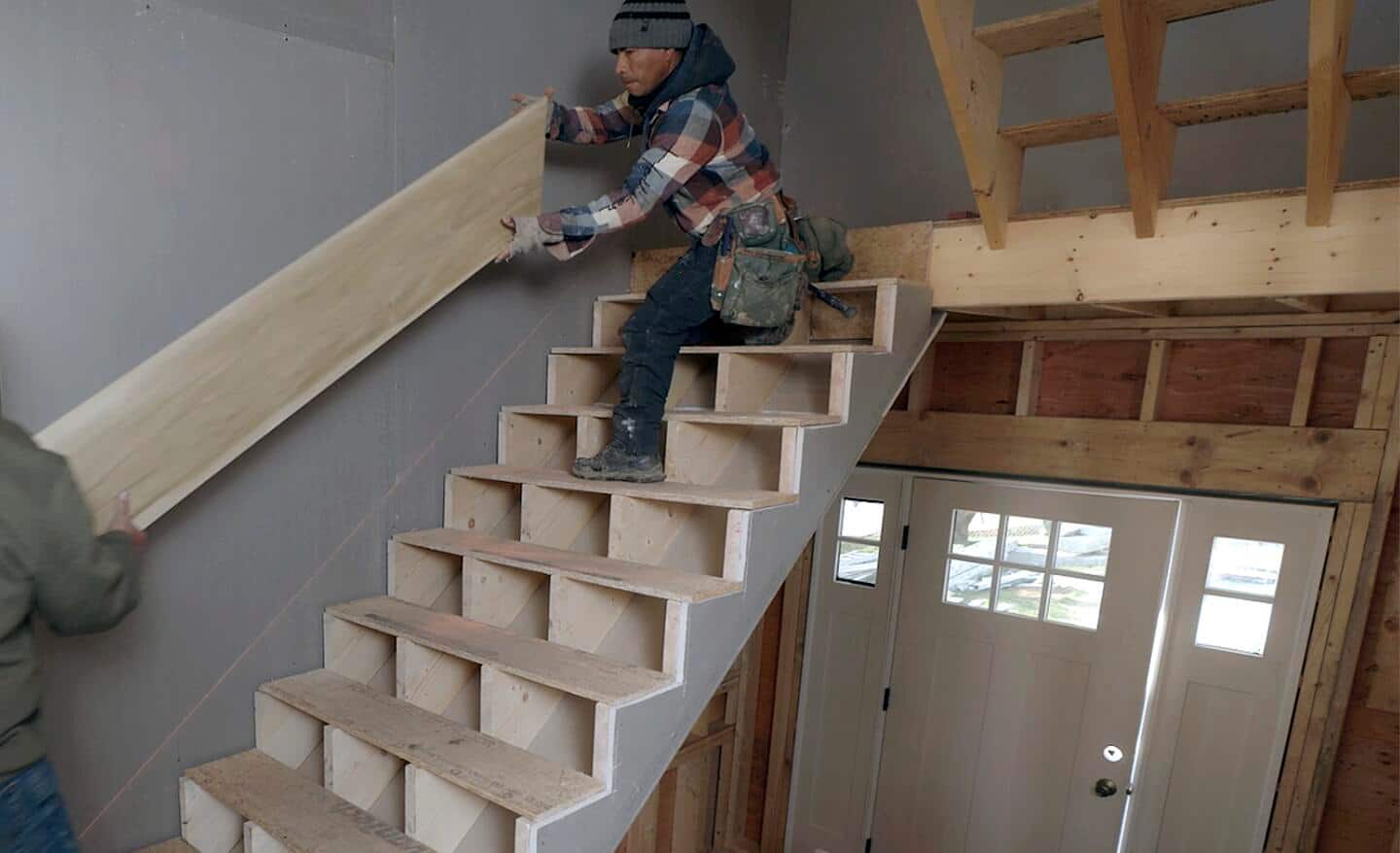 Workers move a 1x12 skirtboard into place to install it in a staircase that is under construction.