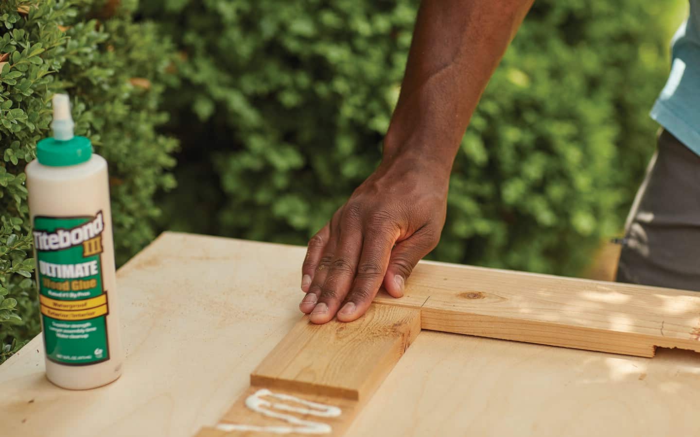 A person holds down a small piece of wood that is being glued to a larger piece on the top part of a cold frame.
