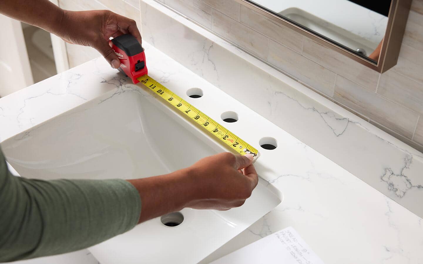 A tape measure is used to find the distance between the mounting holes for a bathroom faucet.