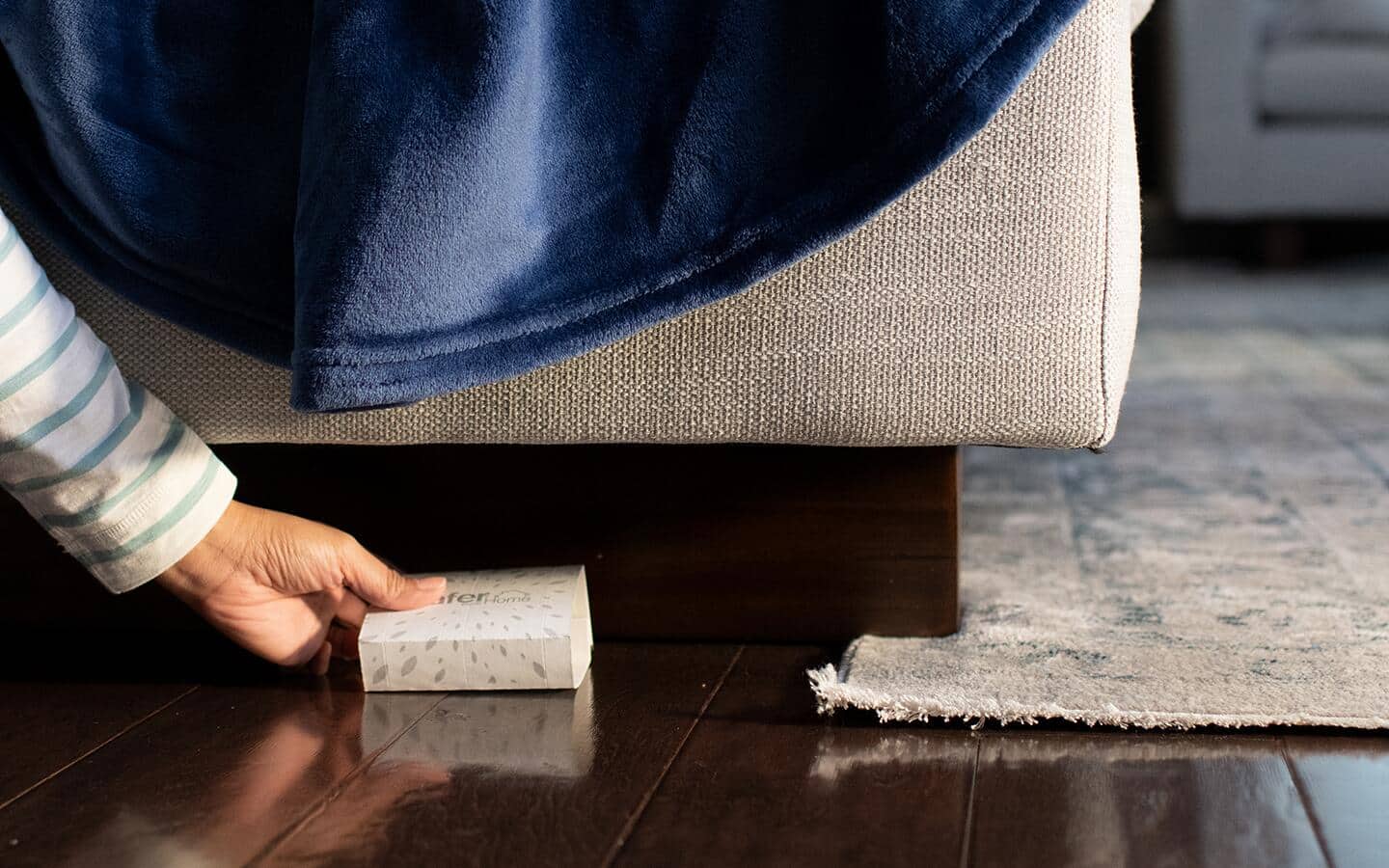 A person places a spider trap under a beige sofa on a wooden floor.