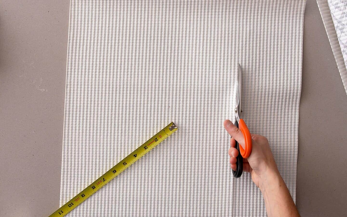 An open tape measure sits on shelf liner as someone cuts it with scissors.