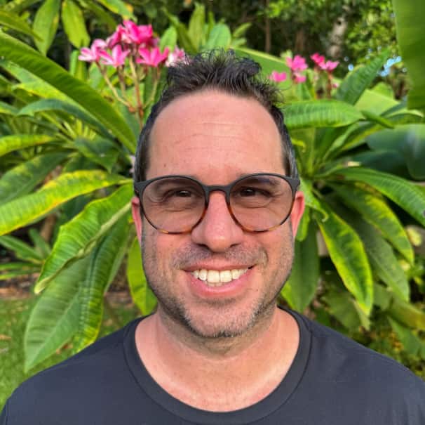Man smiling outdoors with palm trees and flowers behind him.