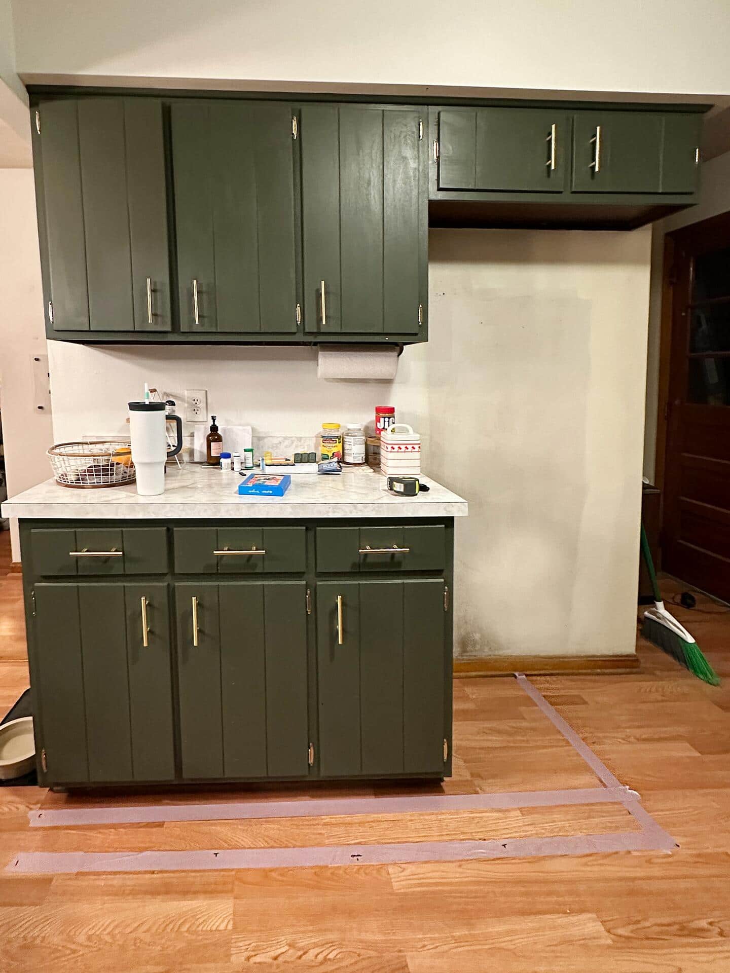  A before photo of Vanessa's kitchen and cabinet spaces, with green cabinetry as they start to make renovations through tape lines on the floor to draw out the new space.