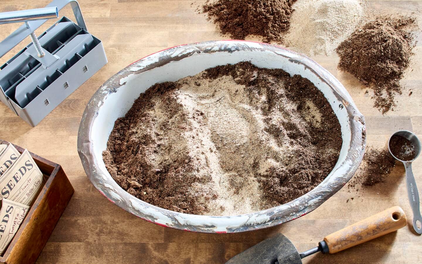 A basin filled with a mix of potting soil and sand for seed starting on a table with supplies like seed packets, tools and a soil blocker