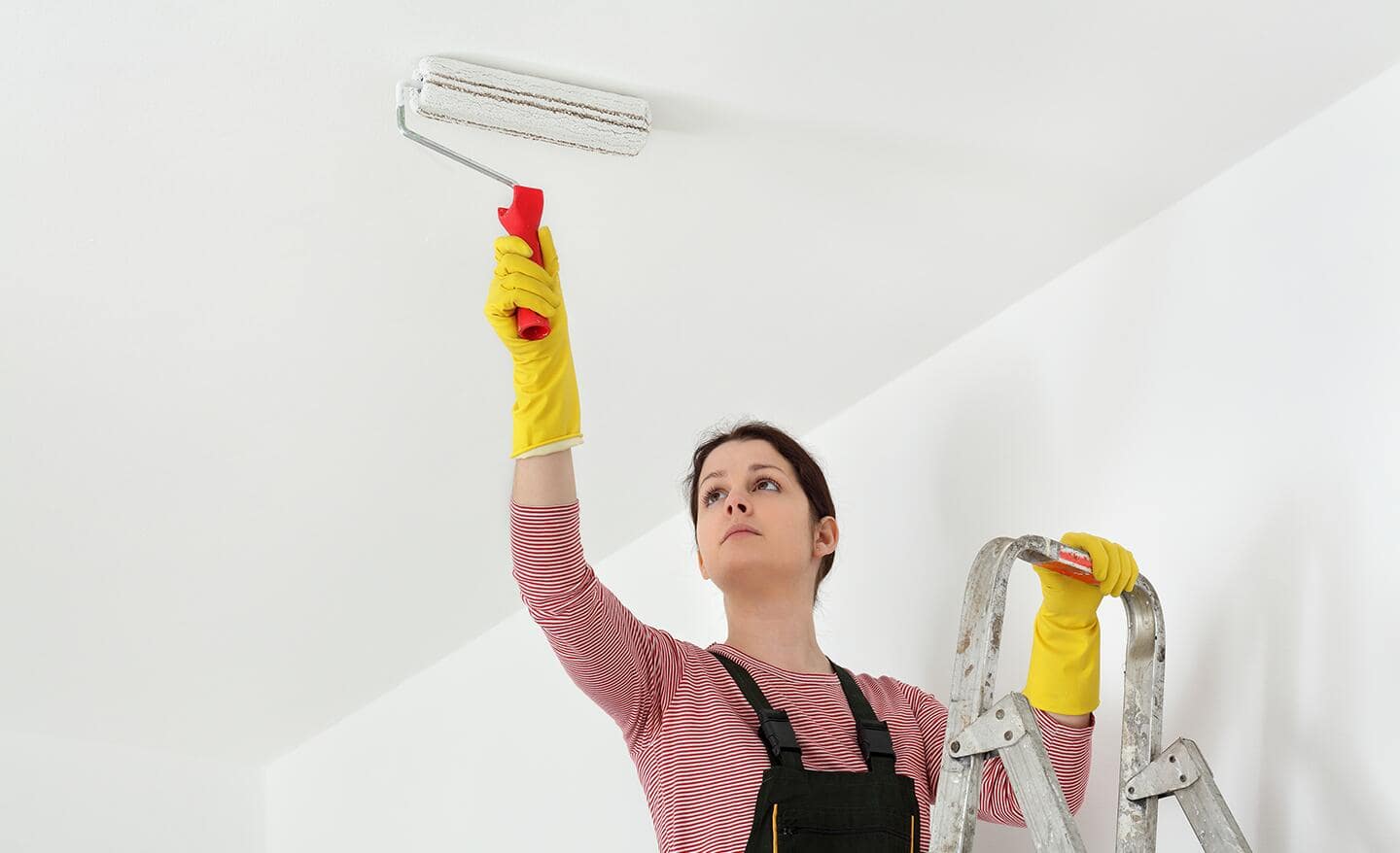 A woman on a ladder uses a paint roller to paint a ceiling. A woman on a ladder uses a paint roller to paint a ceiling.