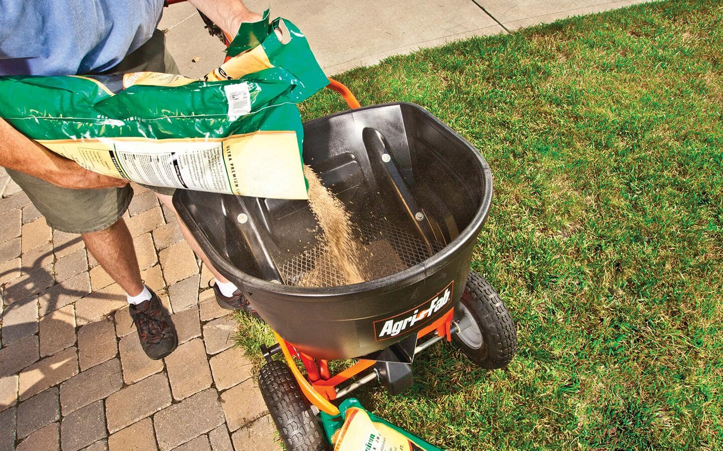 A person pours lawn seed mix into a broadcast spreader. A person pours lawn seed mix into a broadcast spreader.