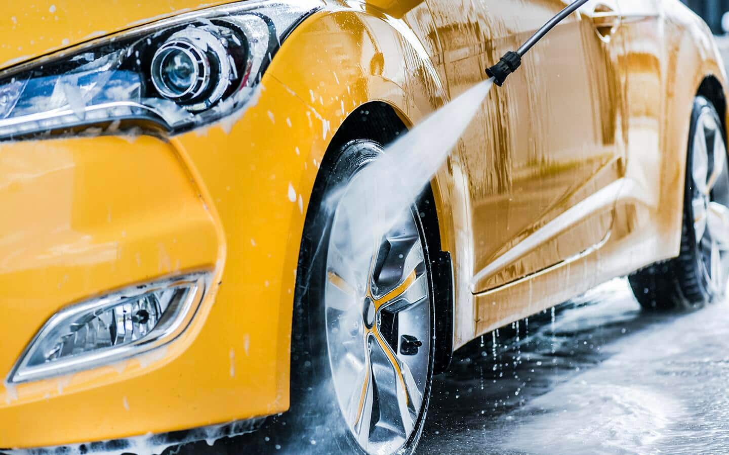 A person washes a tire on a yellow car with the spray nozzle of a hose.