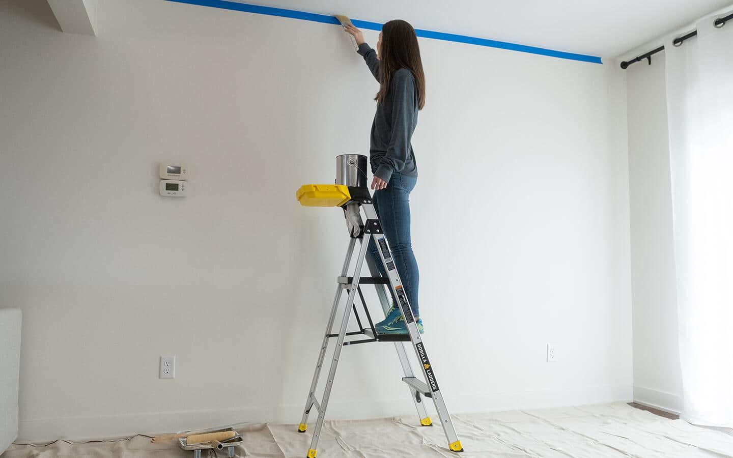 A woman stands on a ladder to paint a ceiling.