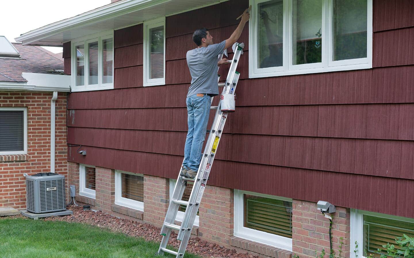 A man uses an extension ladder to reach a window to paint it.