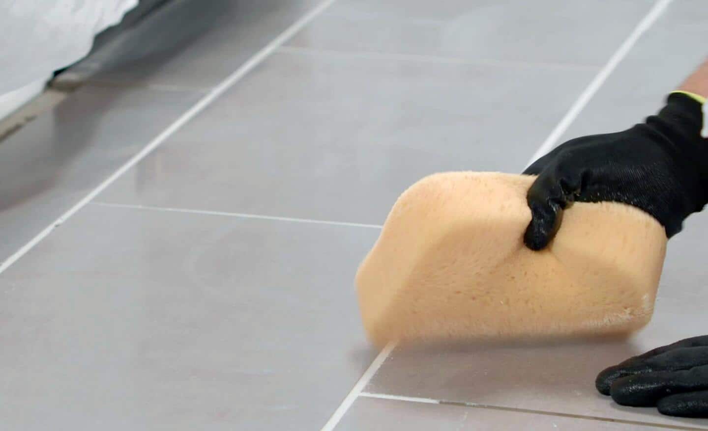 Person uses a damp sponge to wipe grout on a tile floor