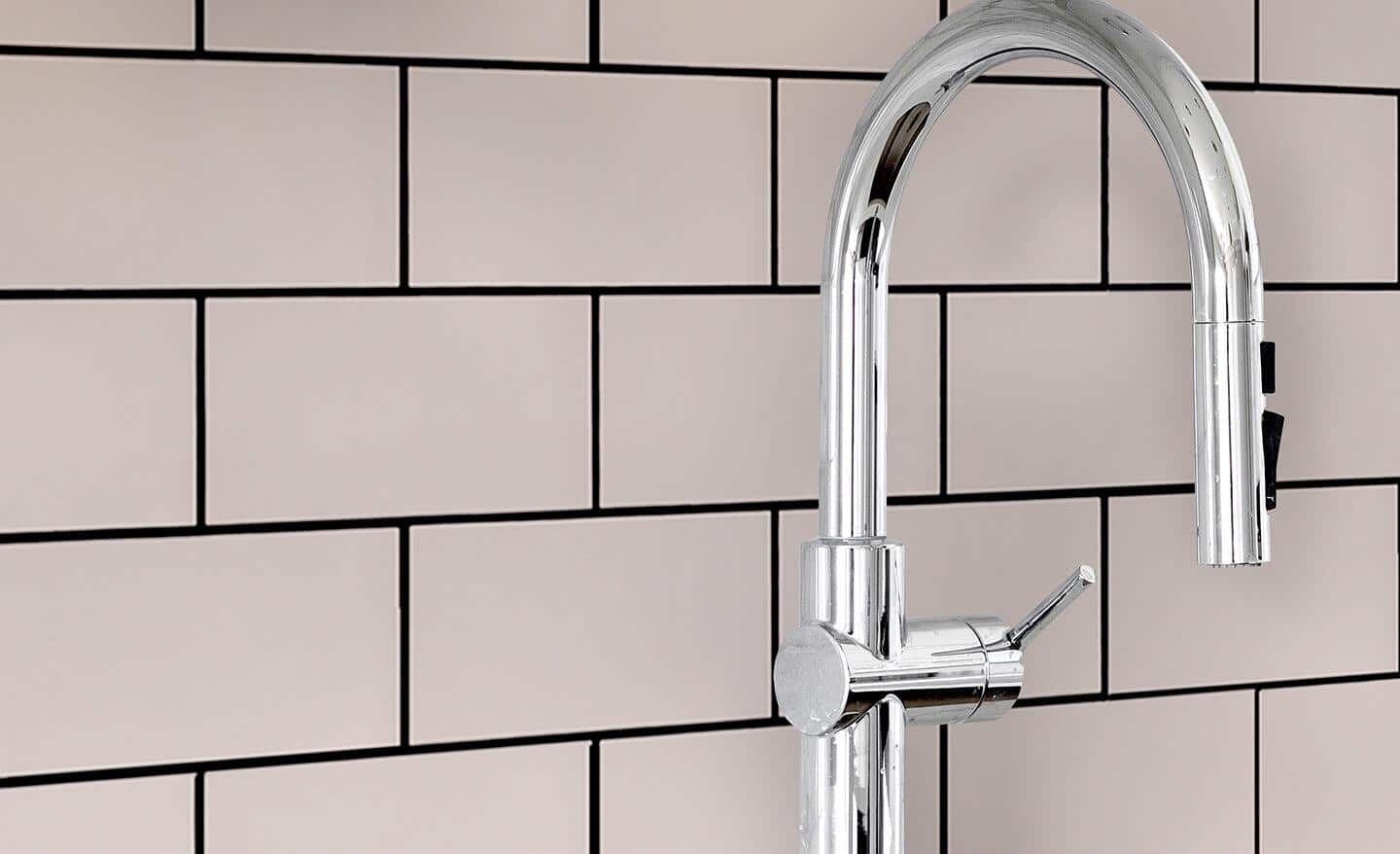 A sink faucet with a backdrop of a light-colored tile wall with dark grout.