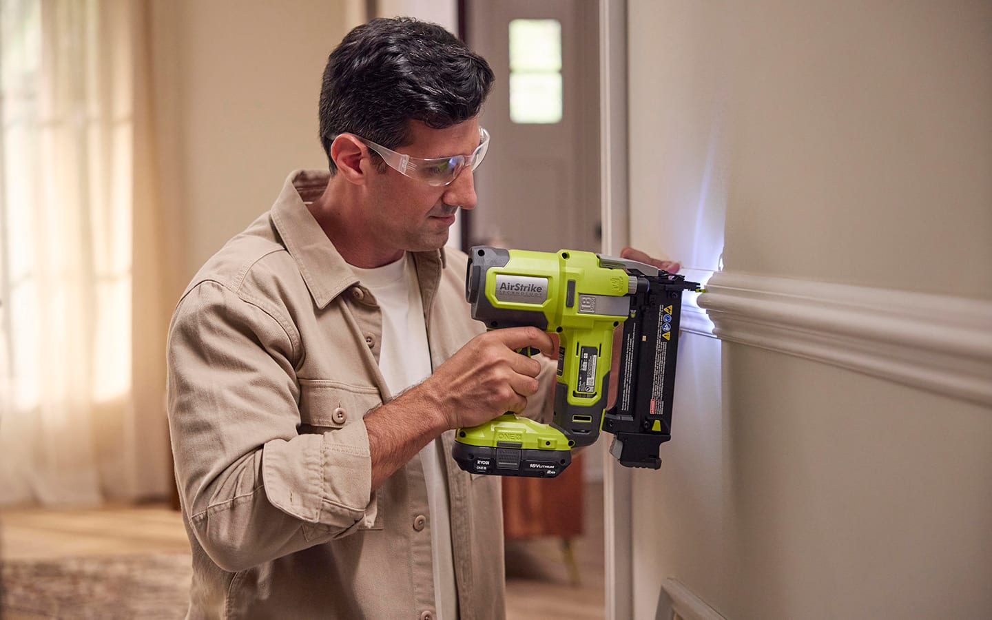 A person in safety glasses attaching moulding to a wall with a cordless nailer. A person in safety glasses attaching moulding to a wall with a cordless nailer.