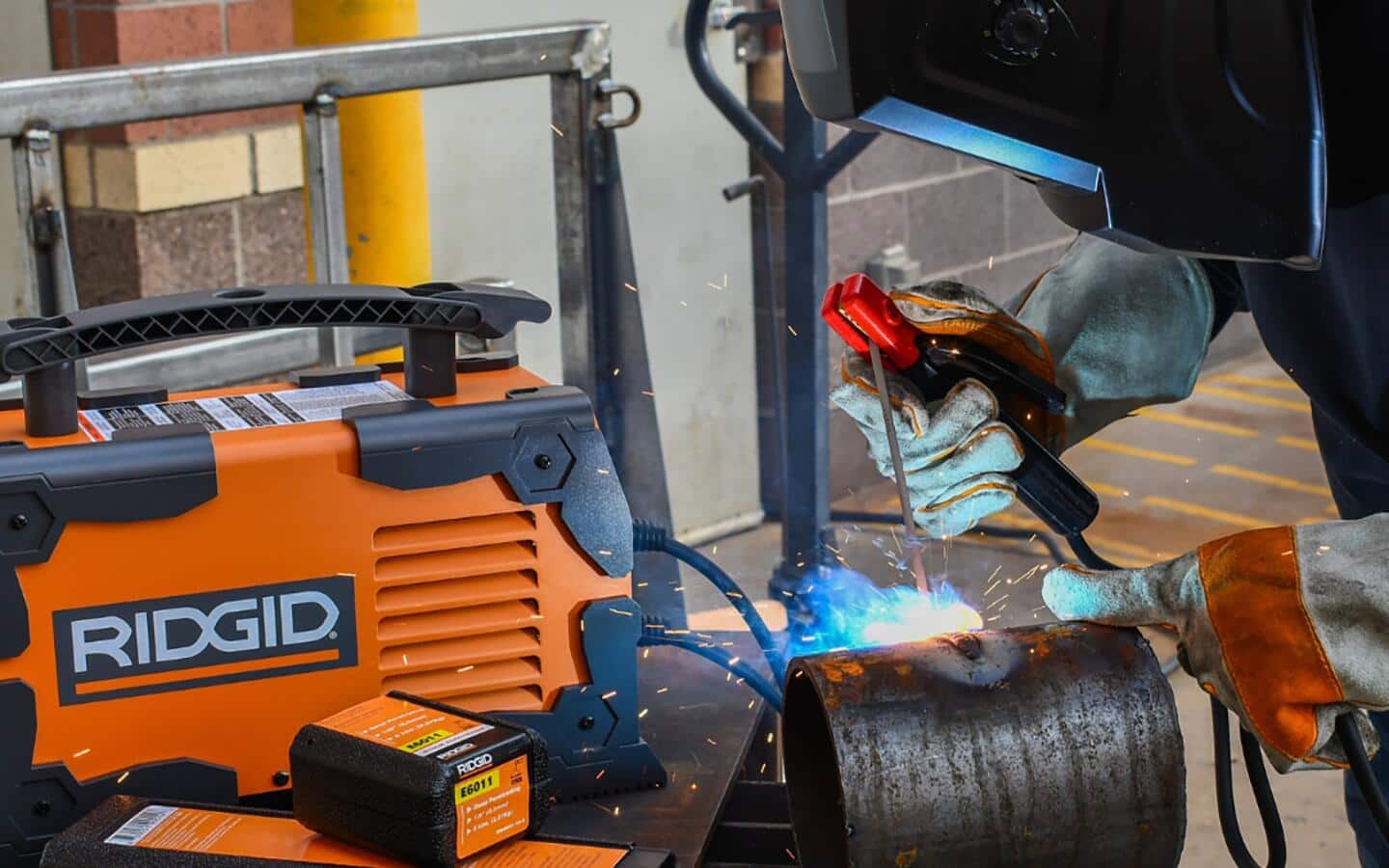 Someone wearing safety glasses and a welding helmet welds a metal cylinder.
