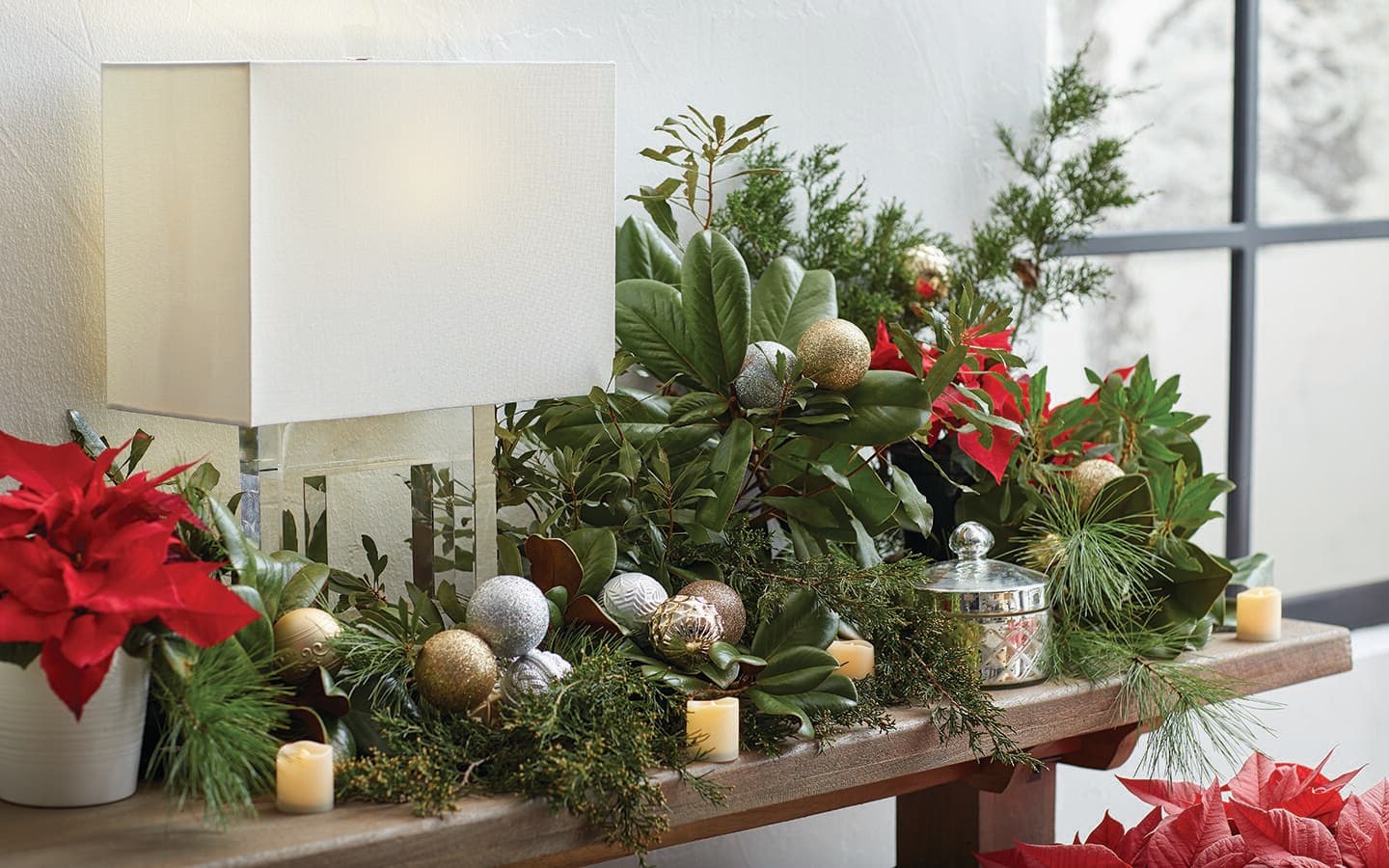 Fresh greenery embellished with metallic Christmas ornaments and candles on a side table.