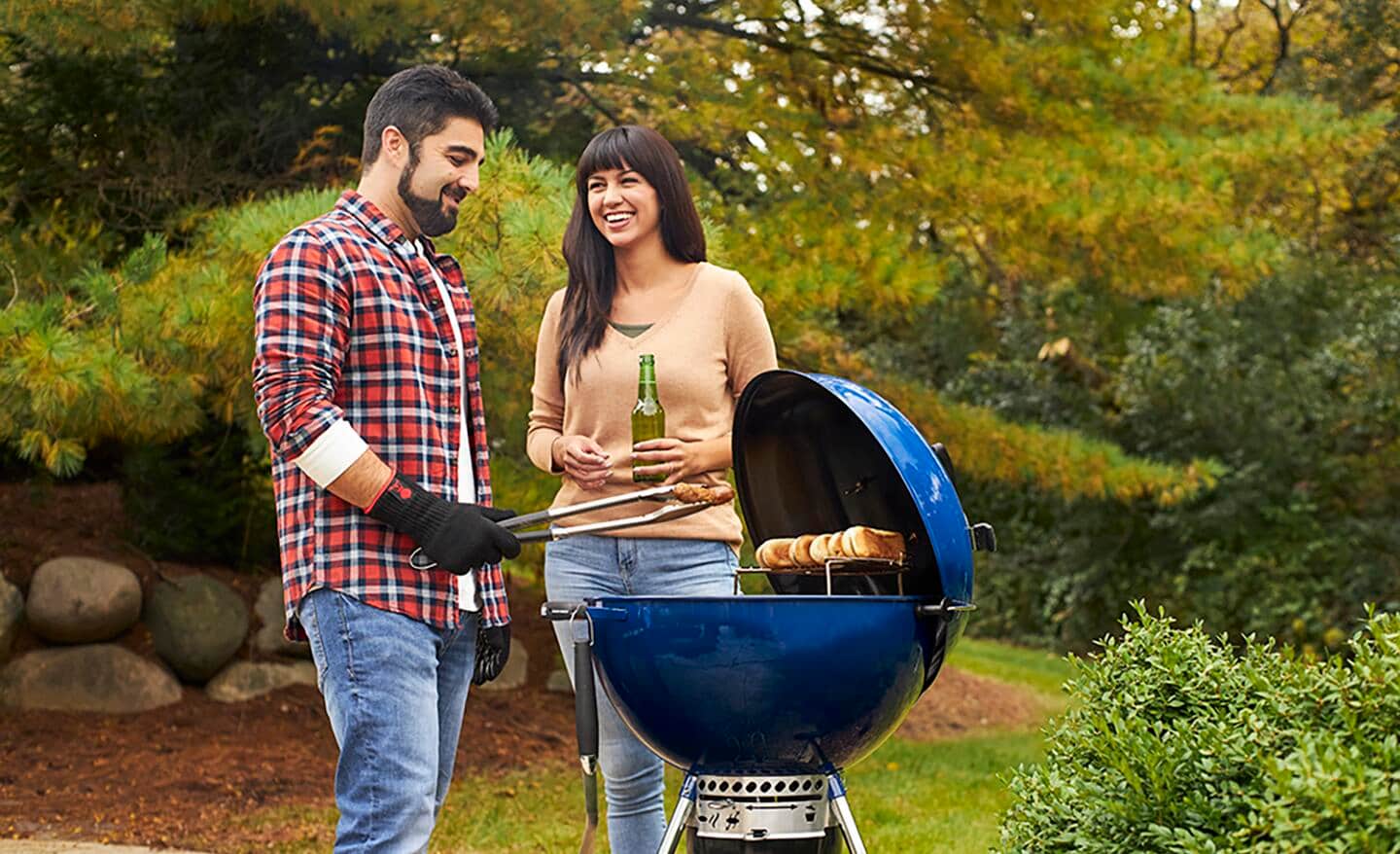A man and a woman cook on a blue charcoal kettle grill on a patio.