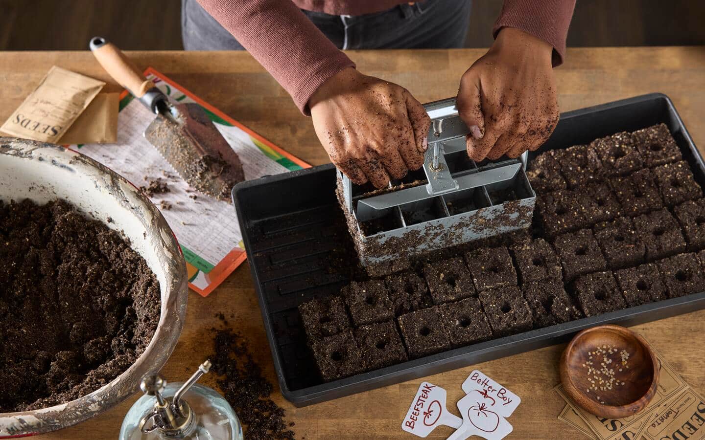 Gardener uses a soil block maker to press out soil blocks in a tray on a table filled with seed starting supplies like tools, seeds and plant markers