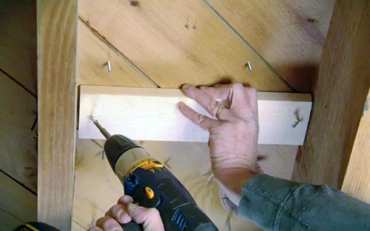 A person drilling into a block of wood to brace squeaky subfloor boards.