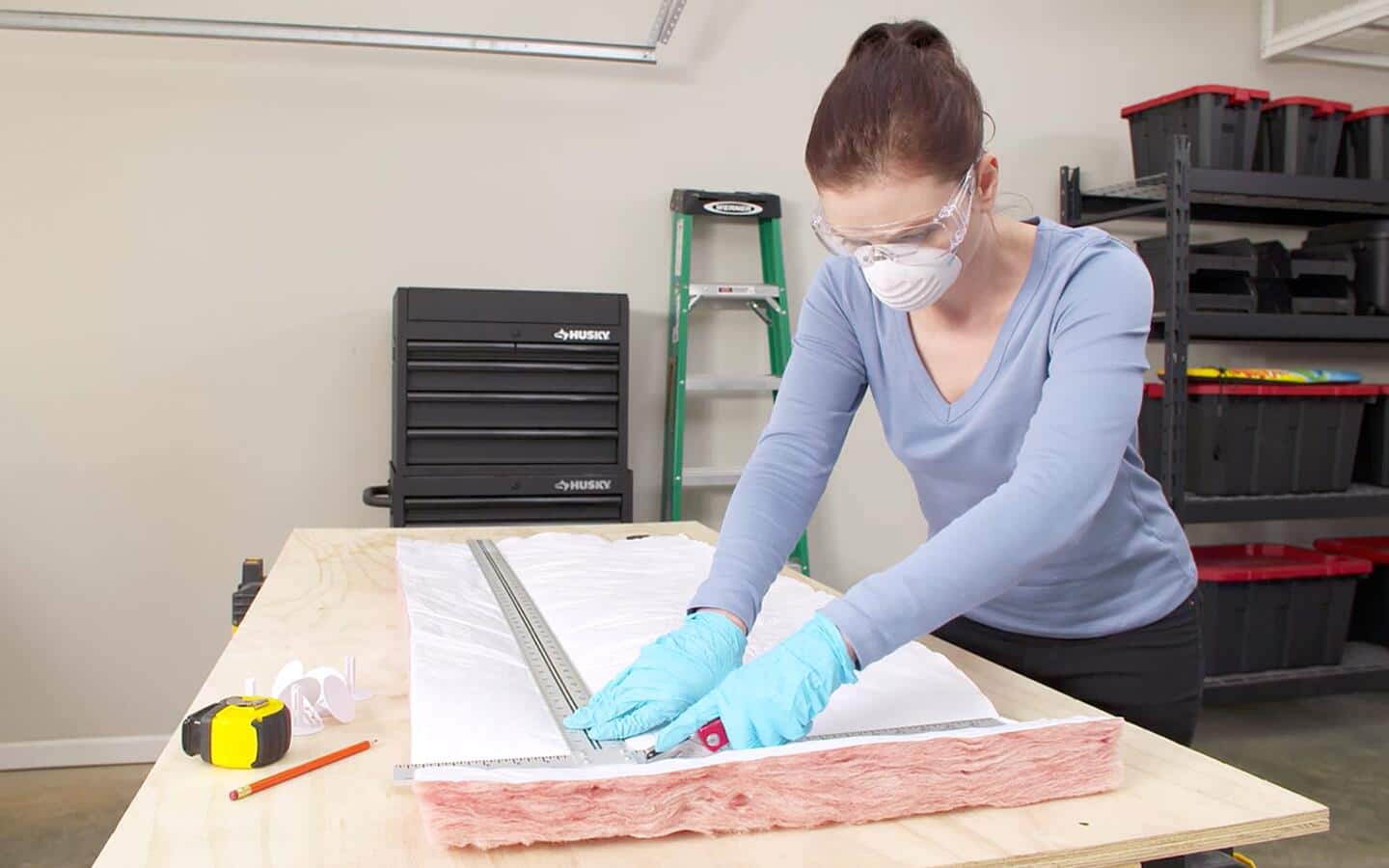A person wearing protective gloves, glasses and a dust mask measuring and cutting fiberglass insulation.