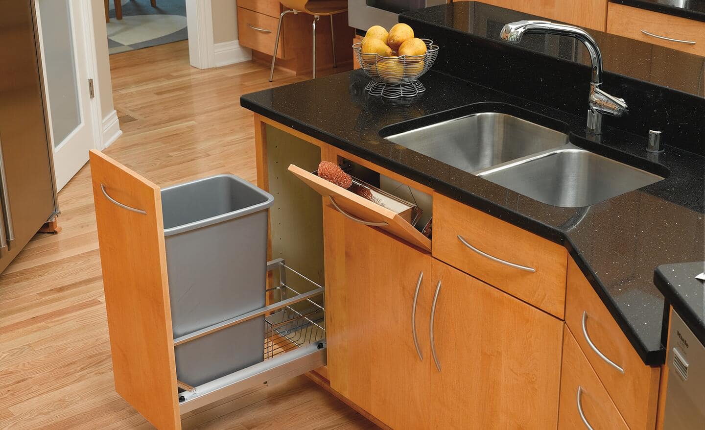 A trash can placed into a pull-out cabinet in a kitchen with light brown cabinets.