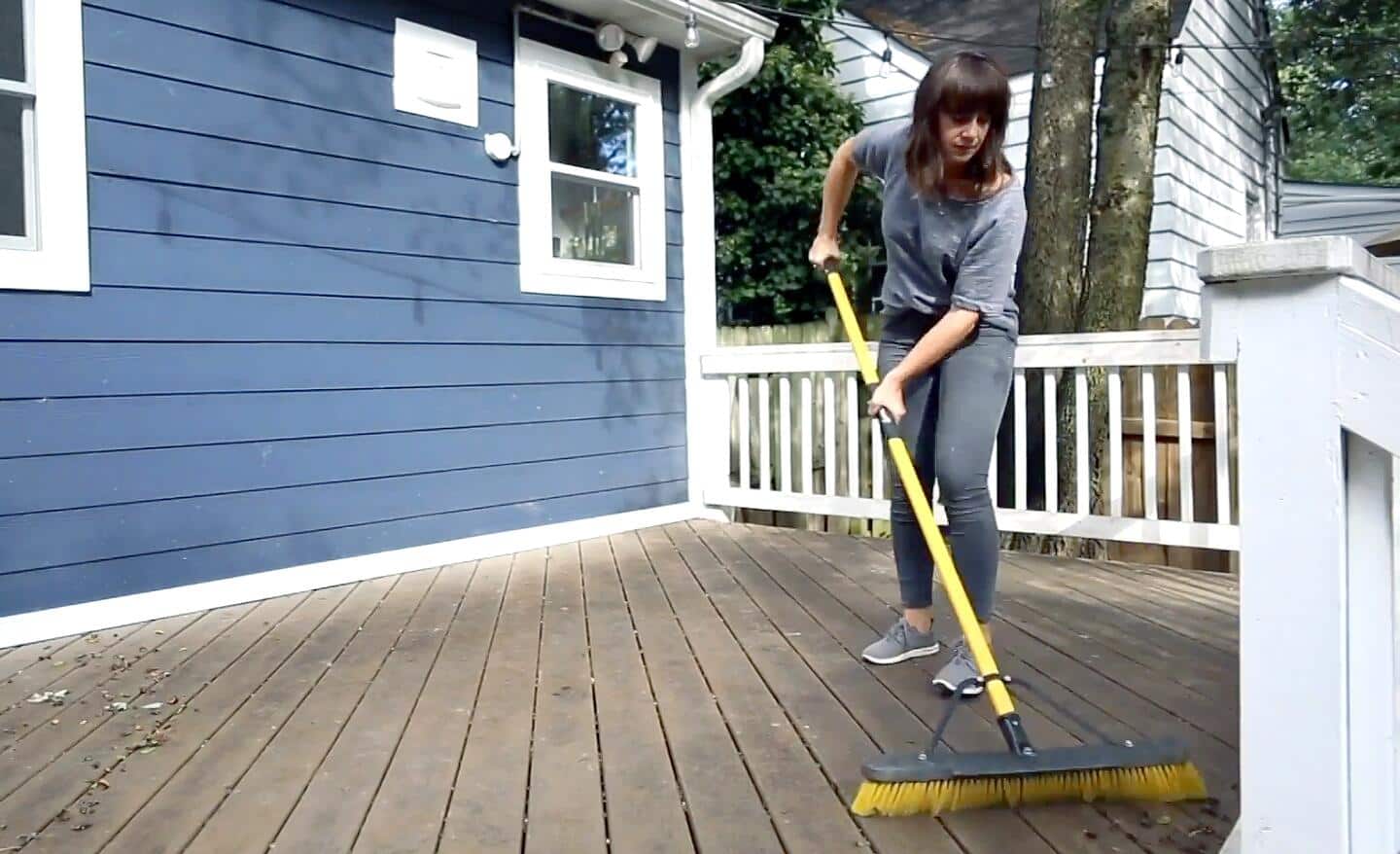 A woman uses a push broom to sweep leaves and other debris from a wooden deck that is to be refinished.