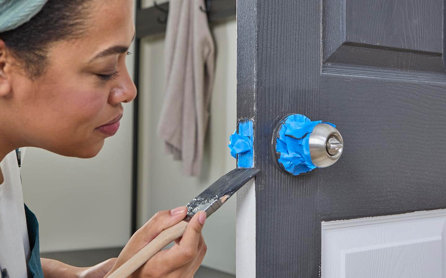 A woman cuts in around a door striker while painting a door that is still attached to the hinges. A woman cuts in around a door striker while painting a door that is still attached to the hinges.