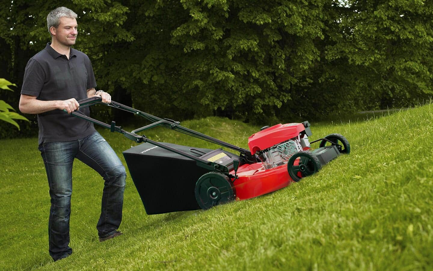 Someone using a red and black push mower on a small, grassy hill. Someone using a red and black push mower on a small, grassy hill.