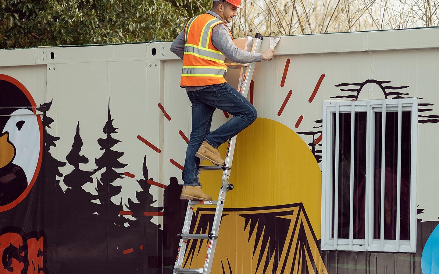 A man wearing high visibility safety gear works from a ladder that is propped against a building.