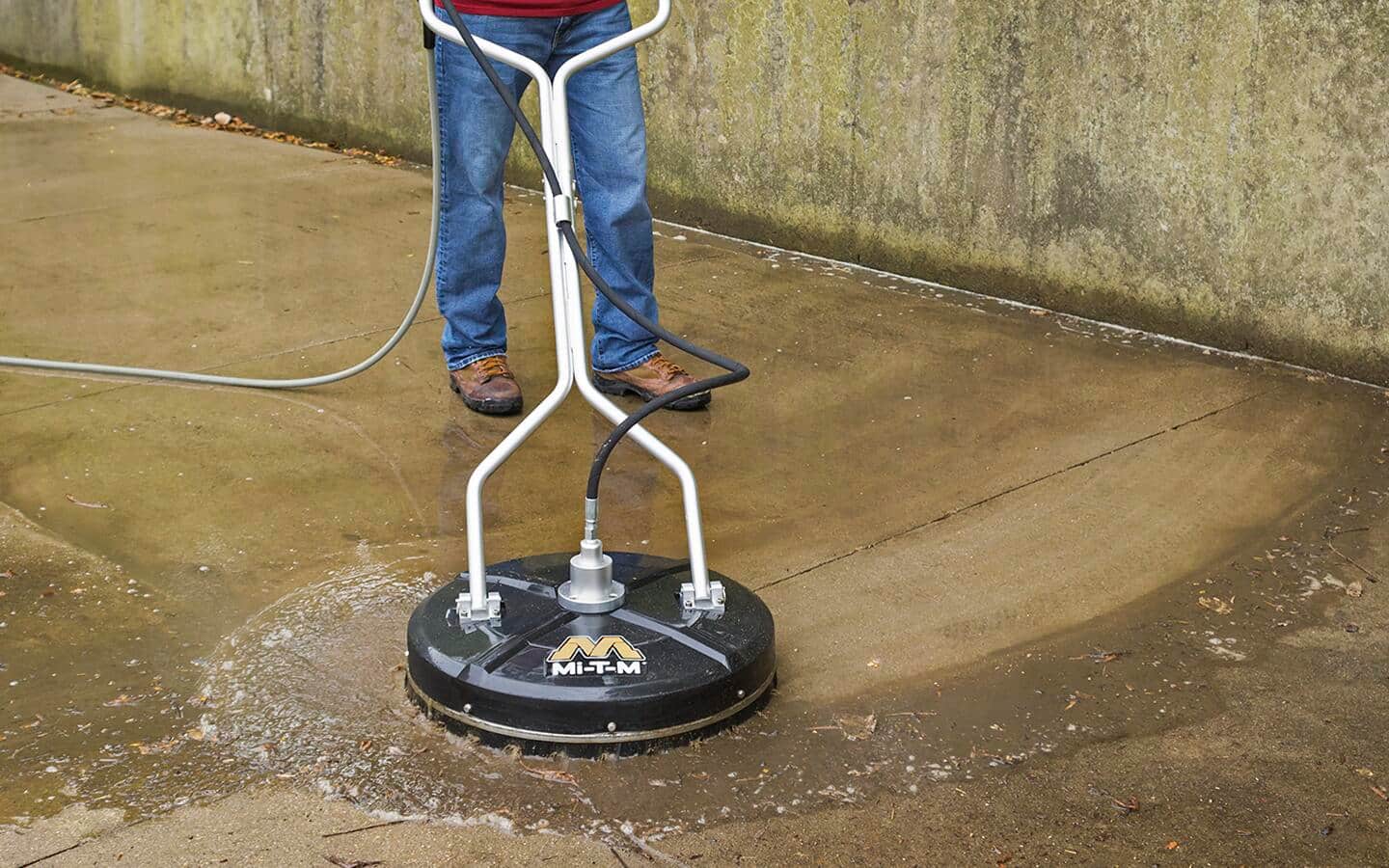 A person using a pressure washer with a deck attachment to clean a concrete walkway