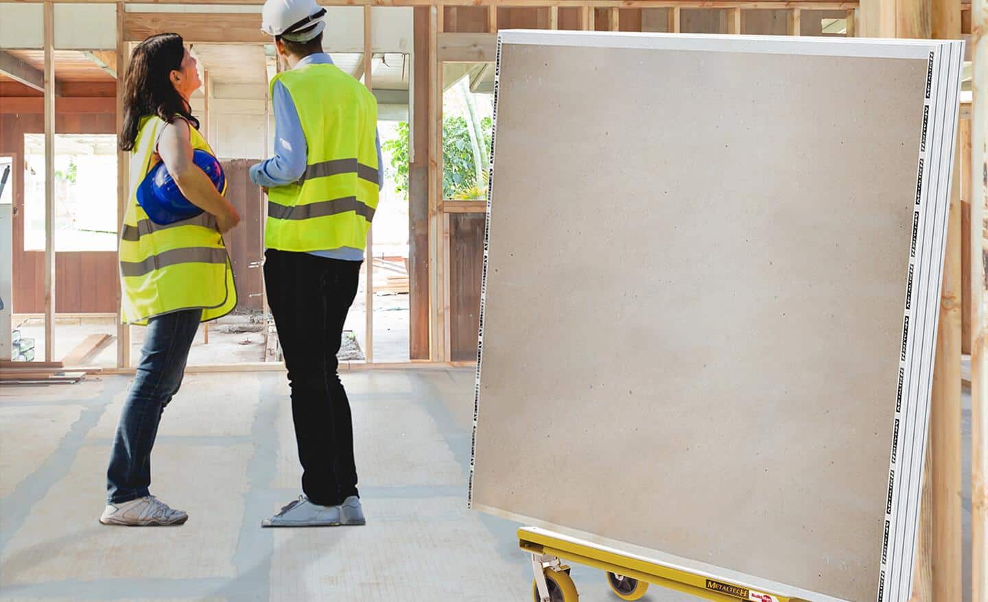Two workers stand next to a stack of drywall sheets propped up in a stand. The workers are looking at the interior frame of the room that has no drywall installed.