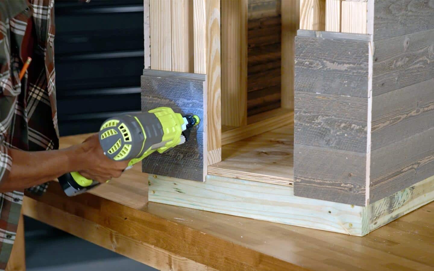 Someone using a cordless nail gun to install siding on the front of a dog house.