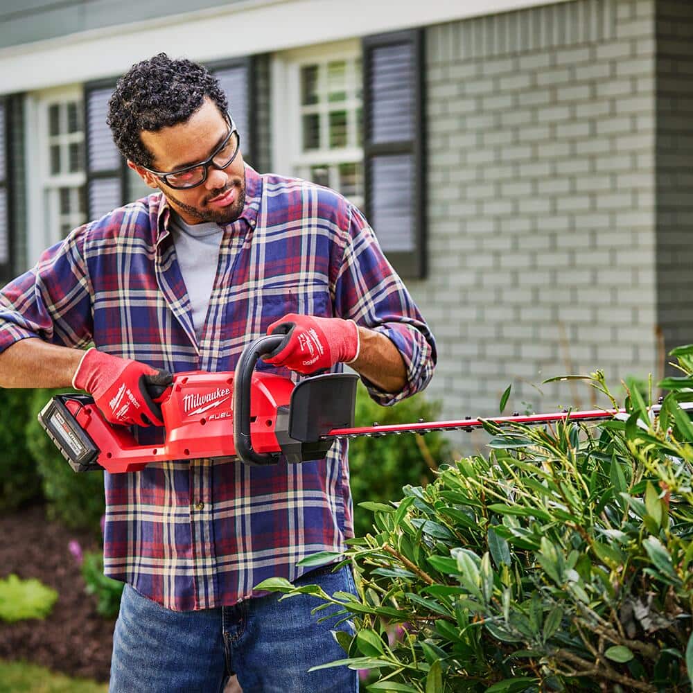 A man uses a battery powered cordless hedge trimmer in front of a brick house.