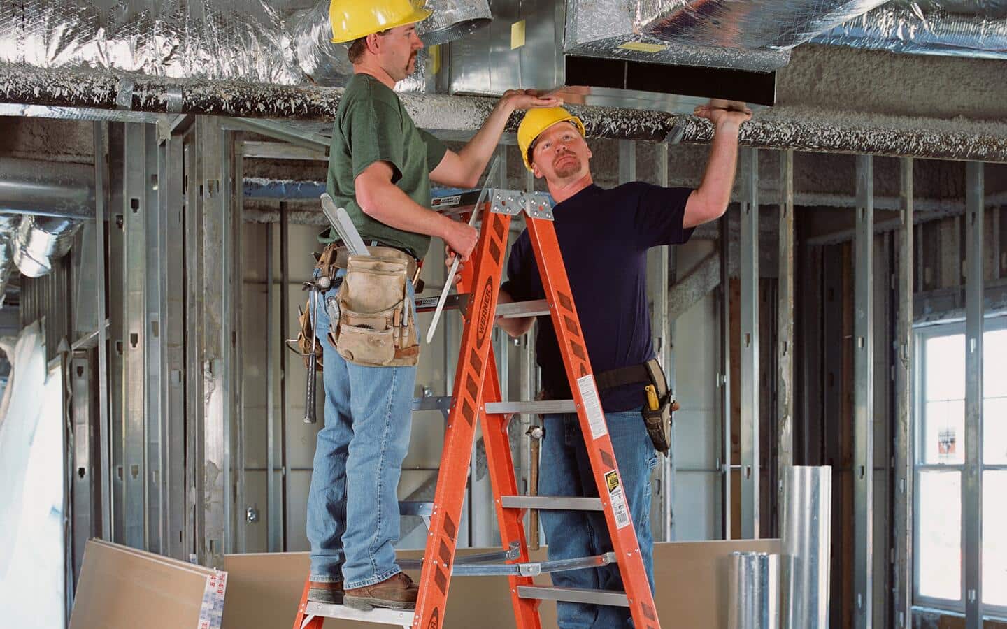 Two people in hard hats on a step ladder working on a ceiling vent.