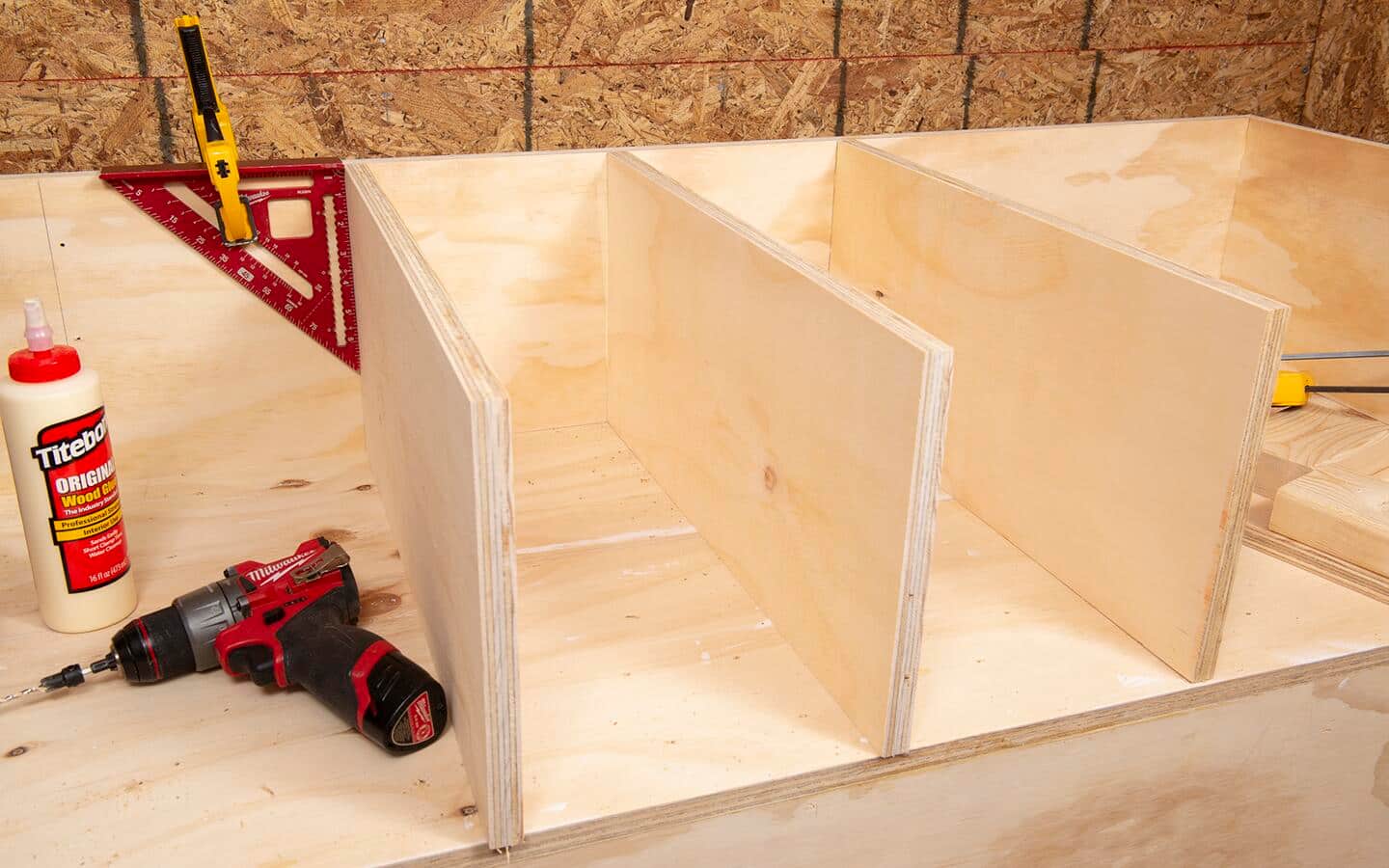 The assembly of a plywood shelving unit with a rafter square, clamp, drill with countersink pilot hole bit and a bottle of wood glue.