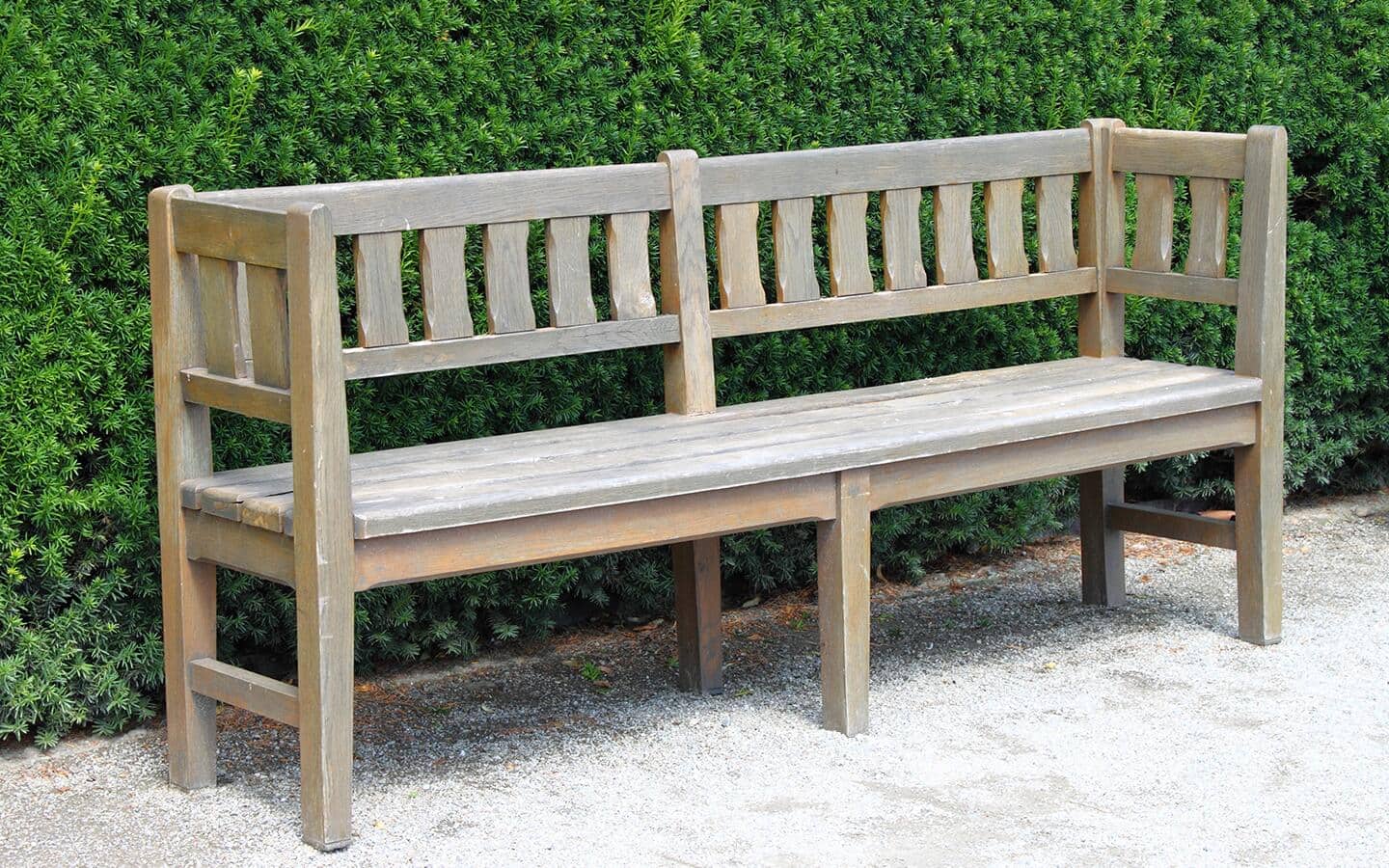 A wood bench sits on gravel in front of a row of bushes.