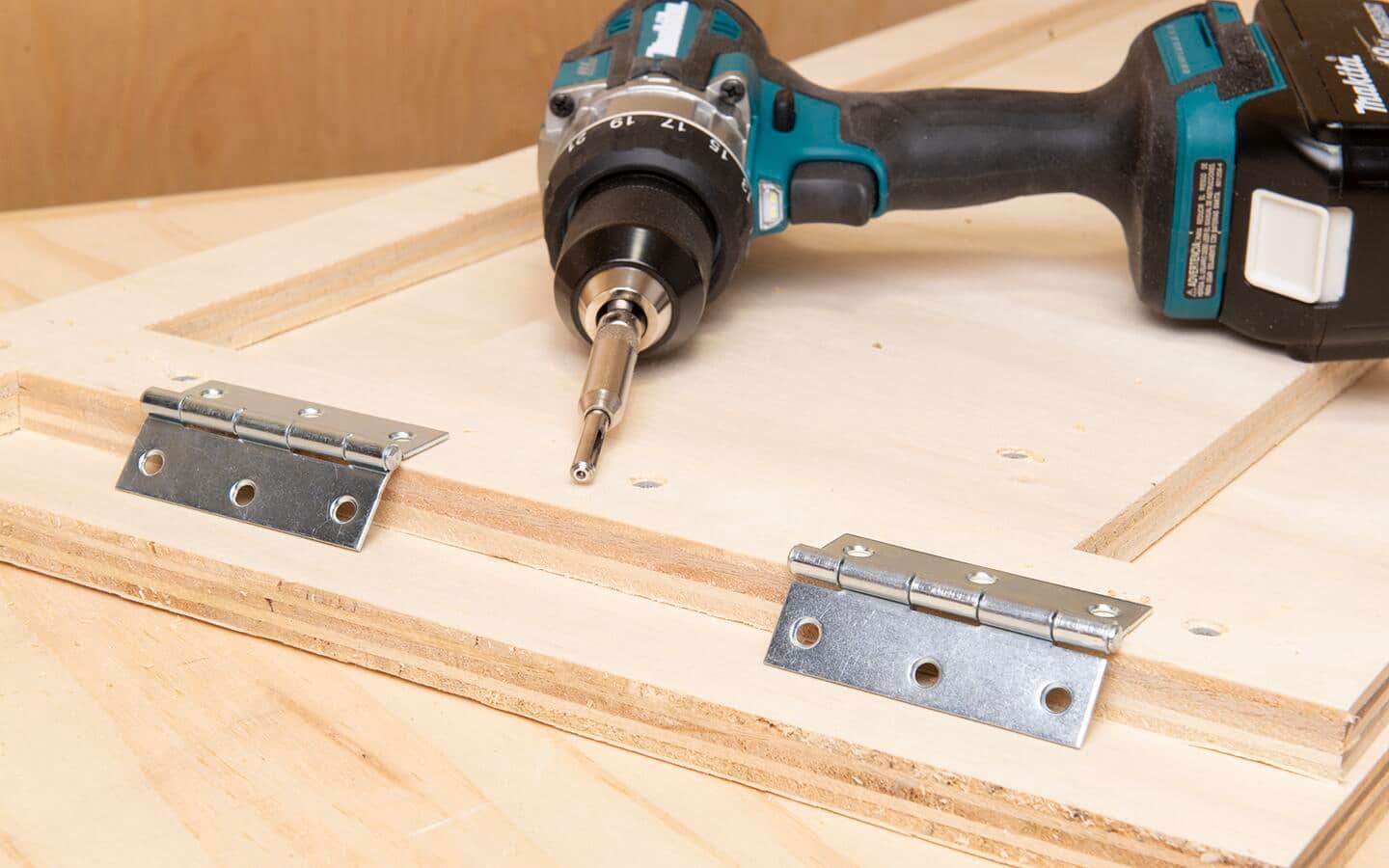 Two hinges are ready to be attached to the hinge boards of a DIY soccer rebounder board, a centering drill bit is chucked in a battery powered drill to make the pilot holes. Two hinges are ready to be attached to the hinge boards of a DIY soccer rebounder board, a centering drill bit is chucked in a battery powered drill to make the pilot holes.