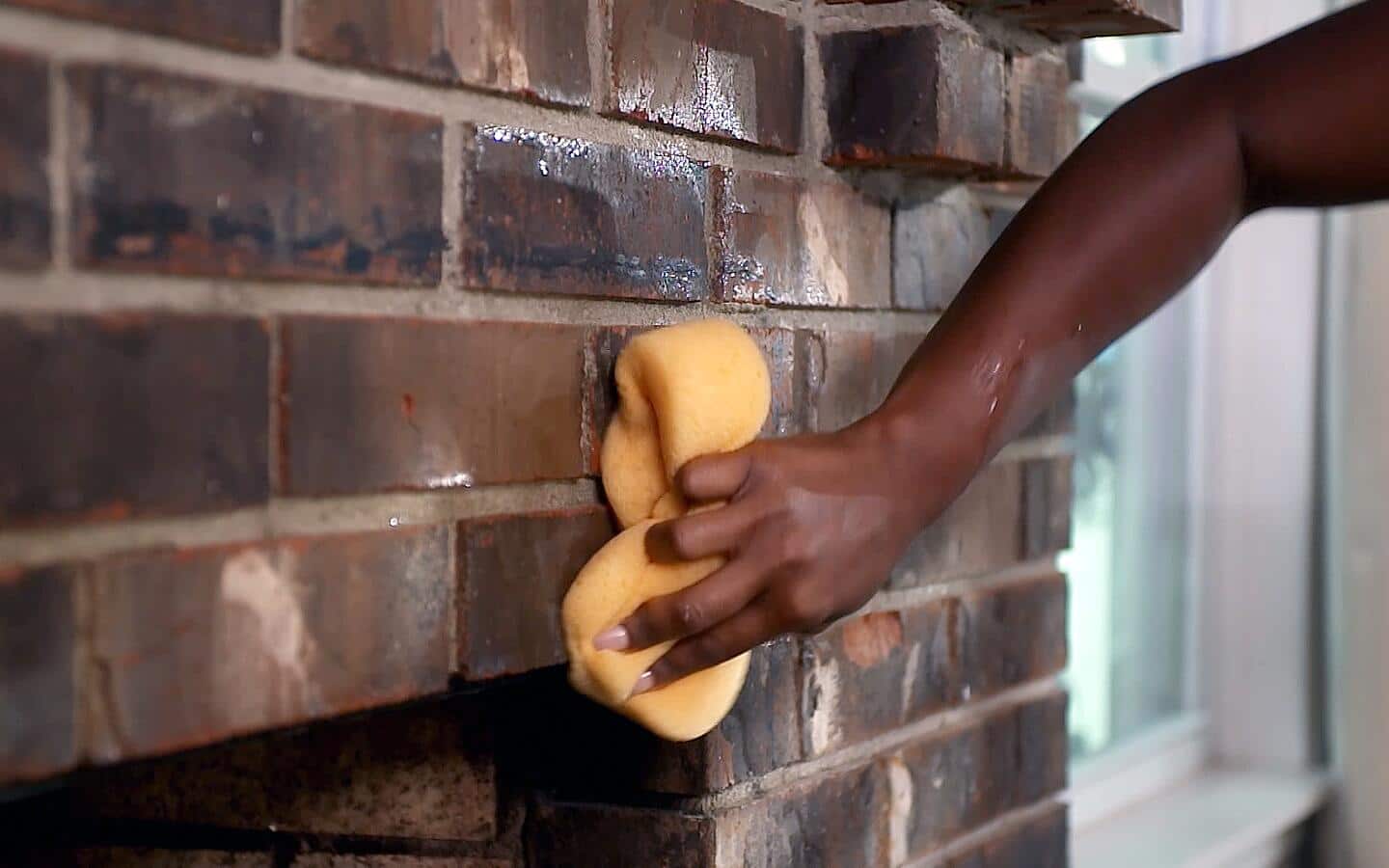 A person uses a sponge to clean dirt from a brick fireplace.