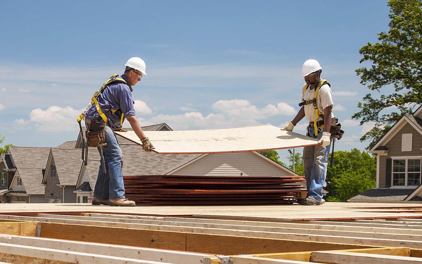 Two workers carrying a sheet of plywood on a roof.