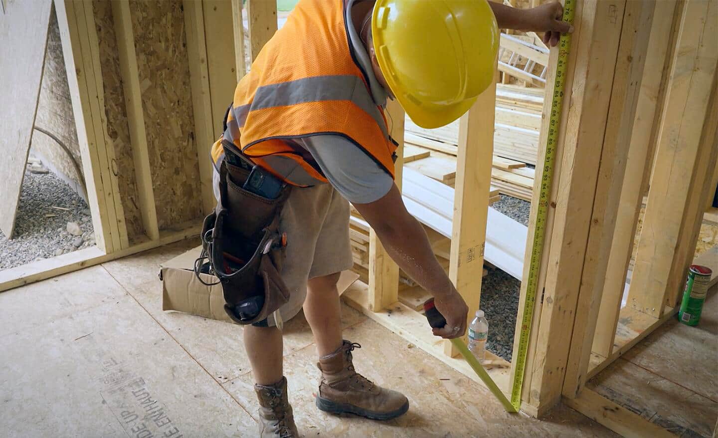 A construction worker, wearing an orange vest and yellow hardhat, measures the framing in a house.