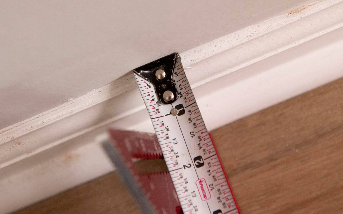 A trim square and a measuring tape are used to find the thickness of previously installed baseboard and shoe molding.