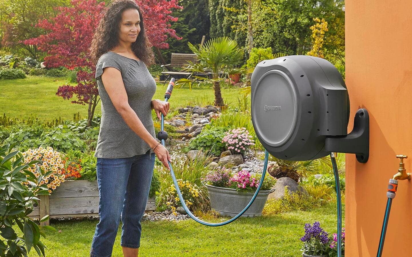 A person pulls a garden hose from an outdoor, wall-mounted garden hose reel.