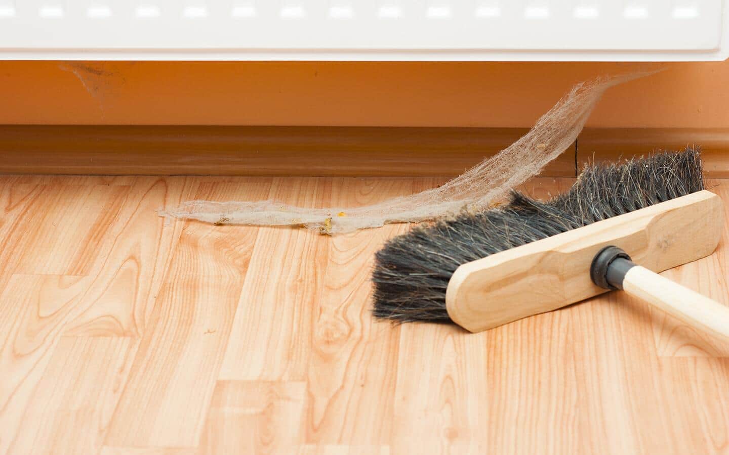 A push broom on a wooden floor in front of a cobweb hanging off the wall.